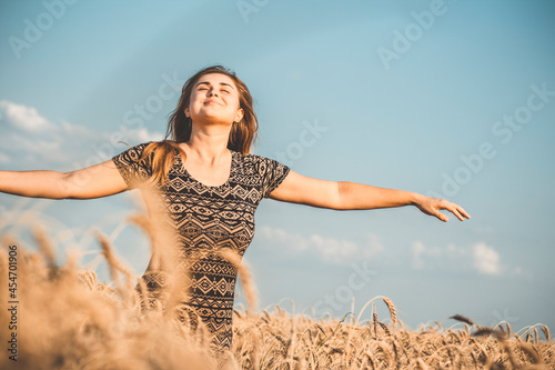 happy beautiful young woman enjoying nature, raising hands on background of cloudy sky in wheat field, girl breathe breathes deeply, freedom and relaxation concept