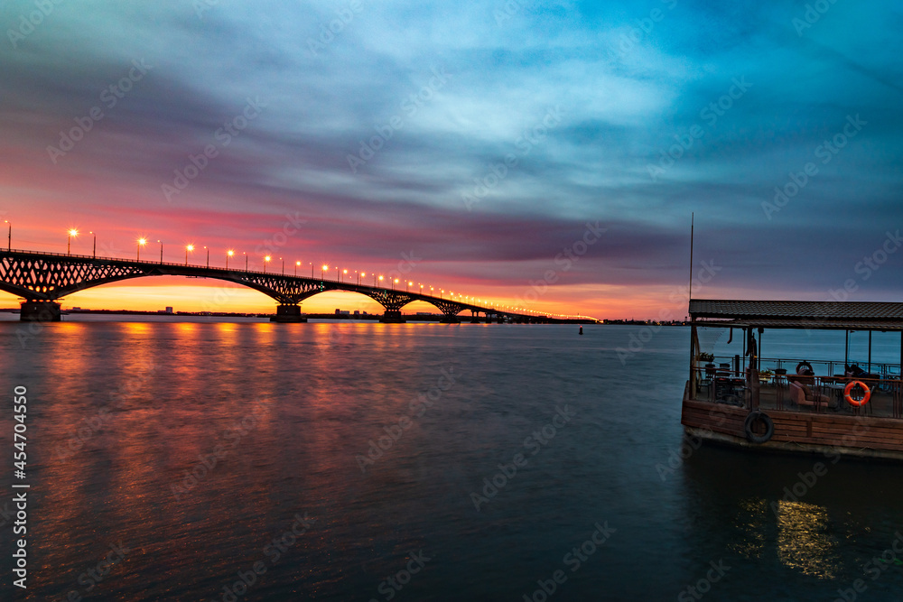 Fototapeta premium Night river before beautiful sunrise. Sunrise on river under dramatic sky. Light on bridge.