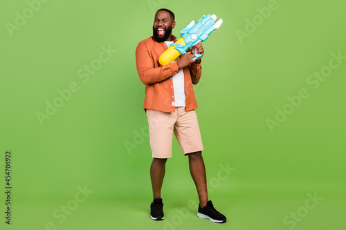 Full size photo of cool young brunet guy hold water gun wear shirt shorts shoes isolated on green background
