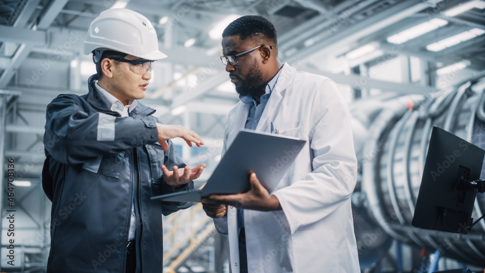 Team of Diverse Professional Heavy Industry Engineers Wearing Safety Uniform and Hard Hat Working on Laptop Computer. African American Technician and Asian Worker Talking on a Meeting in a Factory. Team of Diverse Professional Heavy Industry Engineers Wearing Safety Uniform and Hard Hat Working on Laptop Computer. African American Technician and Asian Worker Talking on a Meeting in a Factory.