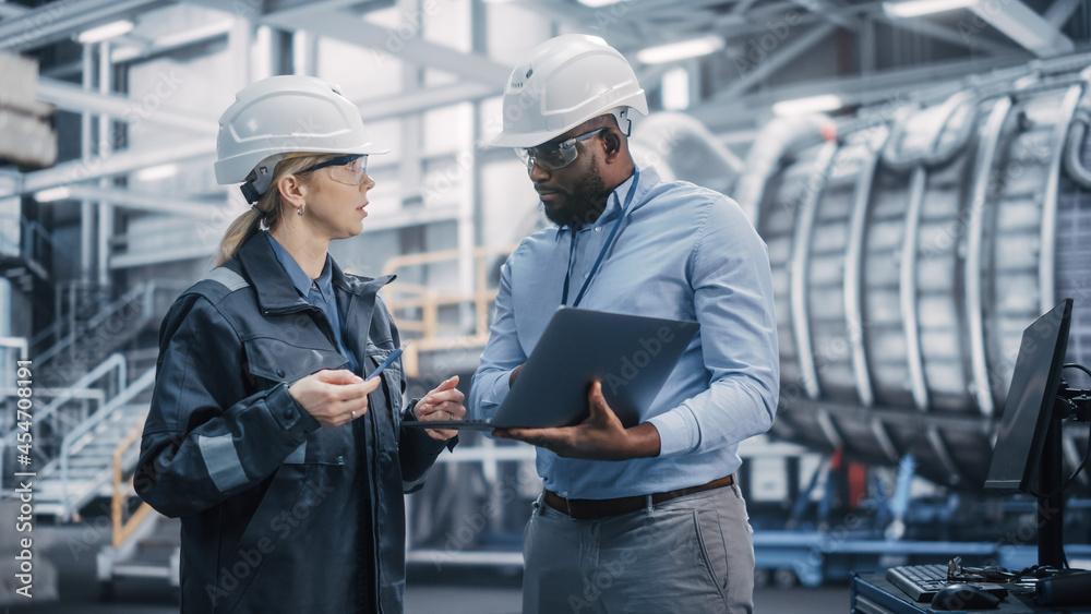 Two Diverse Professional Heavy Industry Engineers Wearing Safety Uniform and Hard Hats Working on Laptop Computer. African American Technician and Female Worker Talking on a Meeting in a Factory. Two Diverse Professional Heavy Industry Engineers Wearing Safety Uniform and Hard Hats Working on Laptop Computer. African American Technician and Female Worker Talking on a Meeting in a Factory.