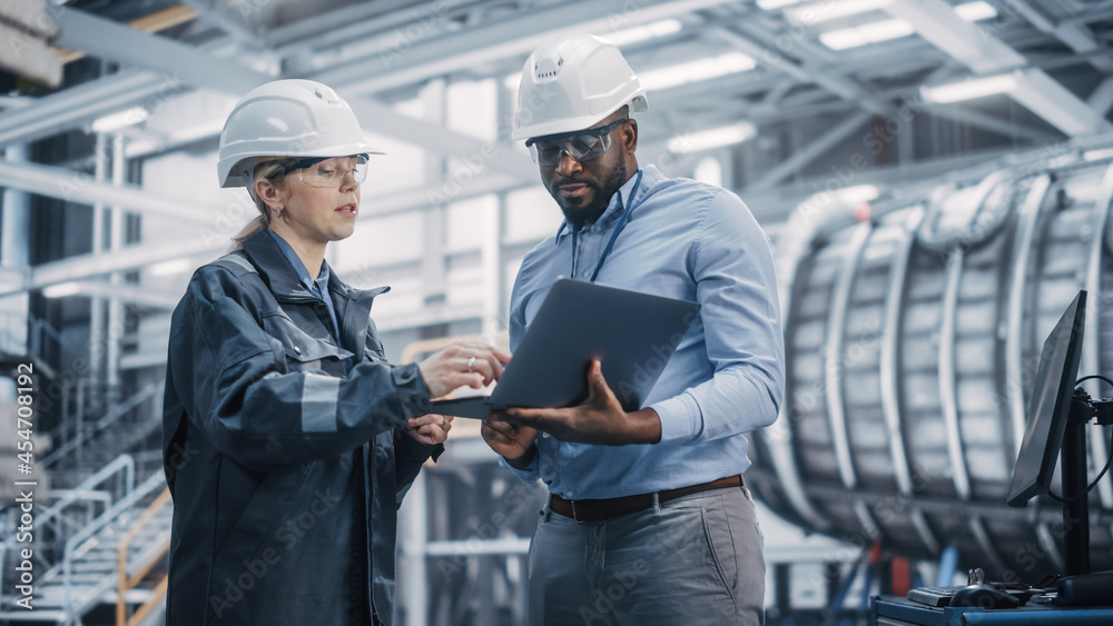 Two Diverse Professional Heavy Industry Engineers Wearing Safety Uniform and Hard Hats Working on Laptop Computer. African American Technician and Female Worker Talking on a Meeting in a Factory. Two Diverse Professional Heavy Industry Engineers Wearing Safety Uniform and Hard Hats Working on Laptop Computer. African American Technician and Female Worker Talking on a Meeting in a Factory.