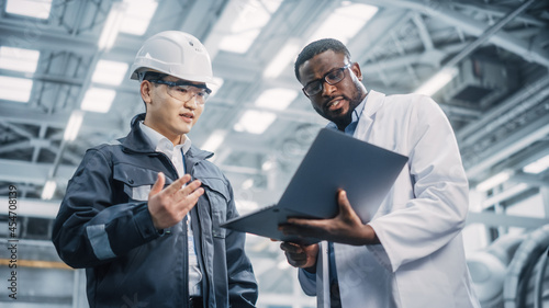 Team of Diverse Professional Heavy Industry Engineers Wearing Safety Uniform and Hard Hat Working on Laptop Computer. African American Technician and Asian Worker Talking on a Meeting in a Factory.