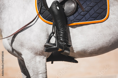 The white horse is wearing a stirrup, a saddlecloth with orange edging and a saddle in which a rider in black boots with spurs sits. Equestrian sports and ammunition. Horse riding.