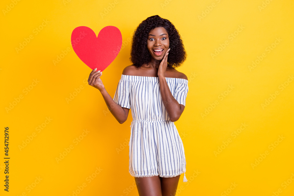 Portrait of attractive amazed cheerful girl holding in hand heart card sign wow isolated over bright yellow color background Portrait of attractive amazed cheerful girl holding in hand heart card sign wow isolated over bright yellow color background