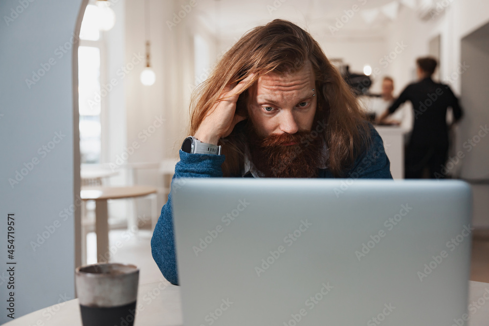 Tired man with red, long hair works at a laptop Tired man with red, long hair works at a laptop