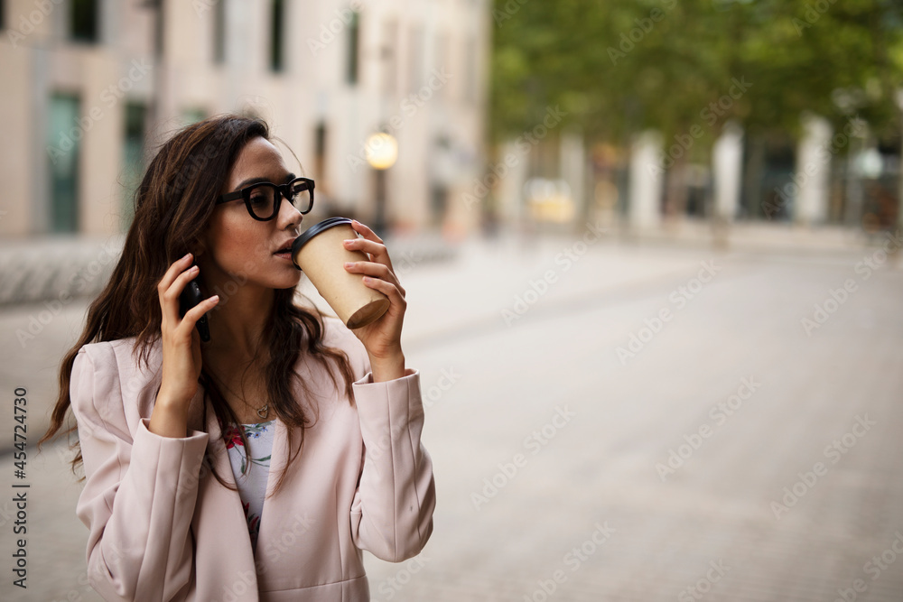 Young businesswoman talking to the phone. Female manager drinking coffee while walking through the city.. Young businesswoman talking to the phone. Female manager drinking coffee while walking through the city..