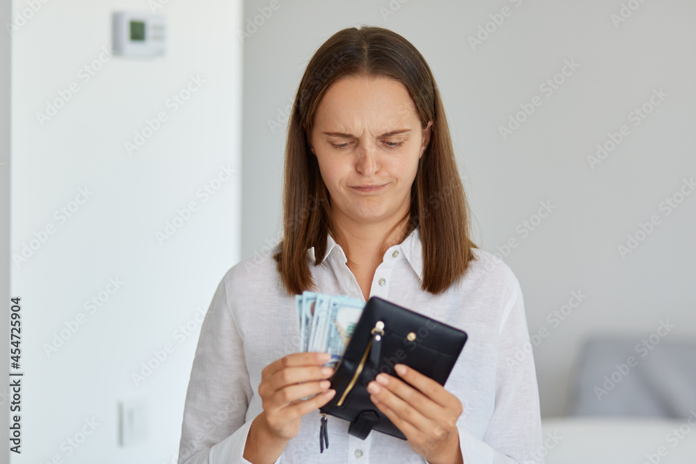 Portrait of upset woman with dark hair wearing white shirt holding in hands wallet with cash, having sad expression, has not enough money for long awaited purchase. Portrait of upset woman with dark hair wearing white shirt holding in hands wallet with cash, having sad expression, has not enough money for long awaited purchase.