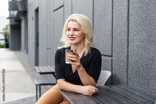 Stylish blonde woman sitting in cafe on terrace drinking coffee, enjoying weekend.