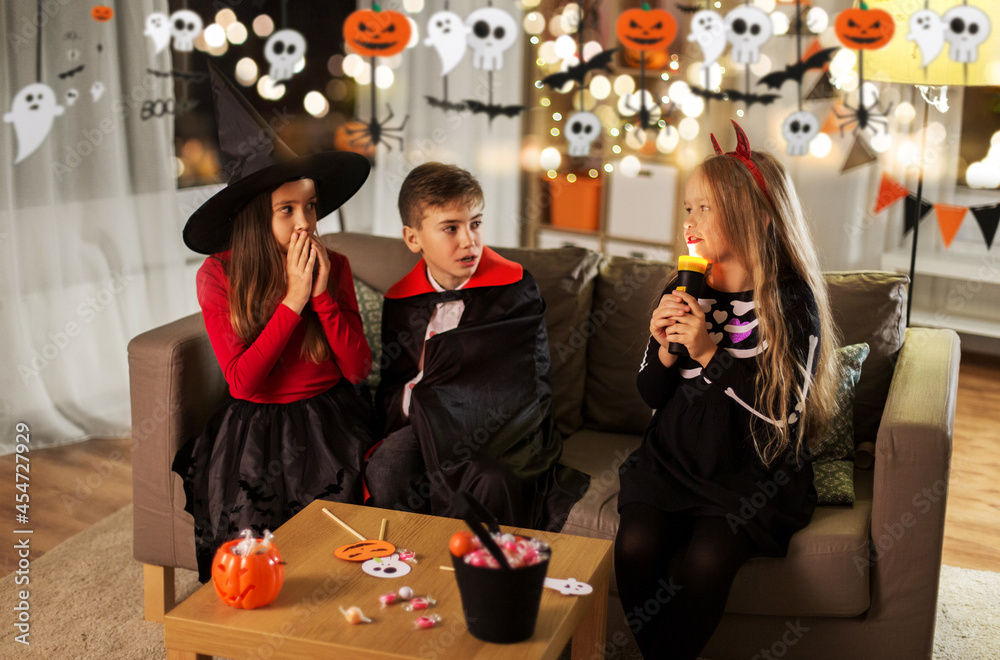 halloween, holiday and childhood concept - smiling boy and girls in party costumes playing with flashlight and scaring each other at home decorated with garland and lights