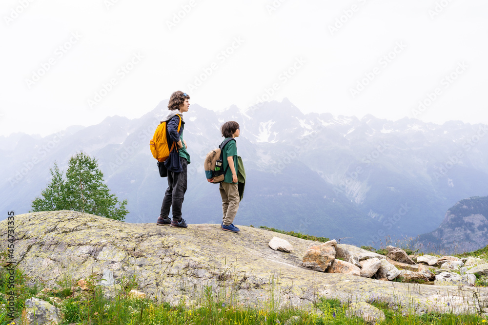 Two boys with backpacks behind them standing on the top of the mountain, looking away, resting after a long ascent. Active vacation concept. Hiking in family.