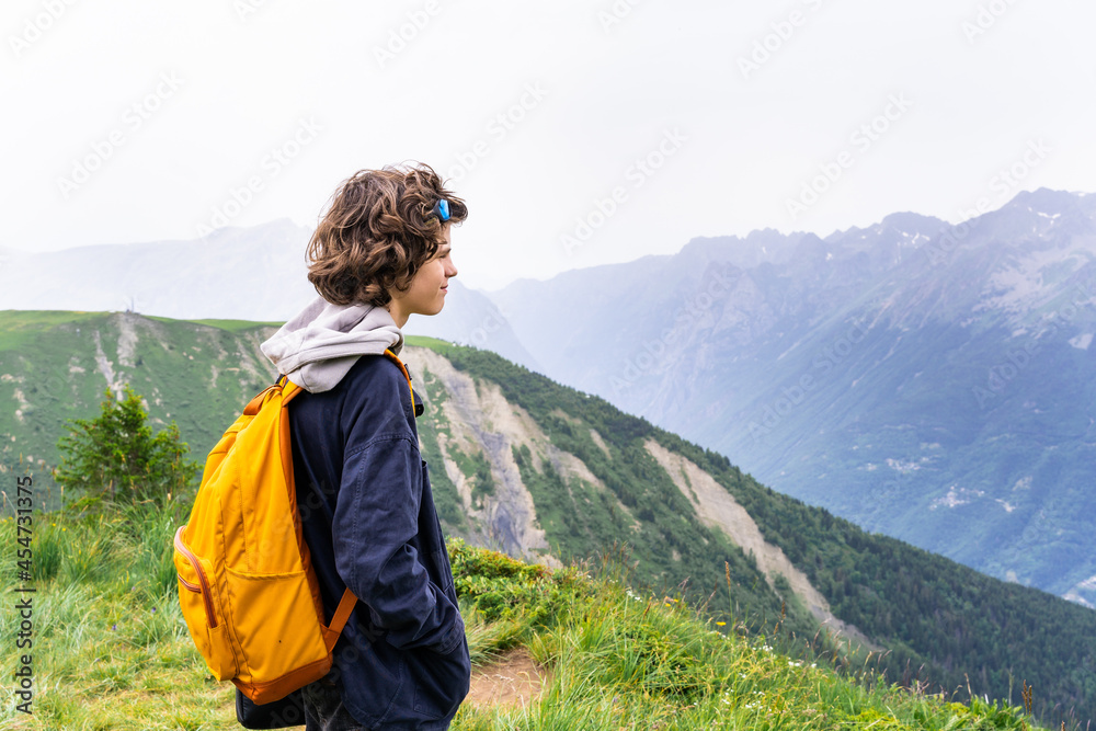 Portrait of a young teenager boy with yellow backpack in a black jacket standing in the mountains in French Alps, looking away with pleasure. Hiking activities in summer.