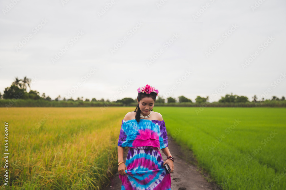 Portrait beautiful Asian woman in a patterned color dress with flowers on her hair, she is touching her dress while walking with color rice field background