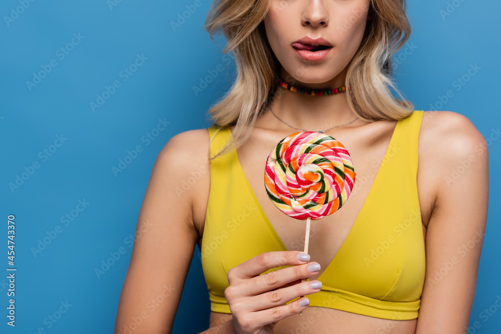 cropped view of young woman in yellow bikini top sticking out tongue and holding sweet lollipop on blue.