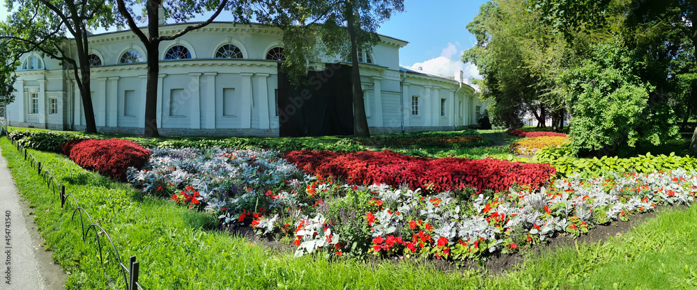 Panorama of a flower garden in a park with a red and green coleus and a pavilion on a sunny summer day.