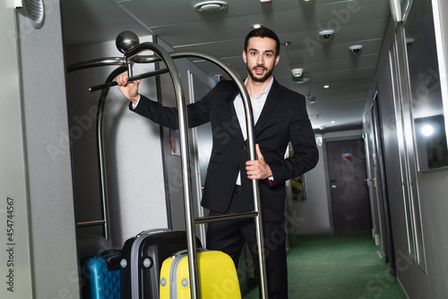 bearded bellboy near metallic bell cart with luggage in hall of hotel photo