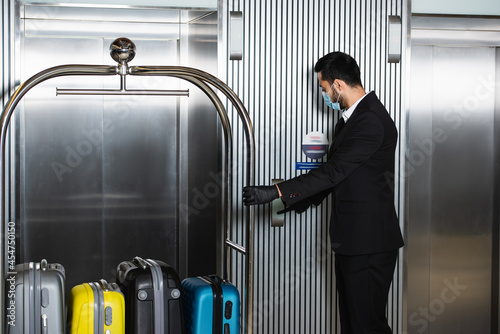 Fotografija bellboy in medical mask standing with bell cart near elevator in hotel