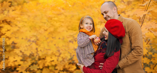 A happy laughing family with a little daughter in their arms against the background of autumn orange foliage of maple trees. A child is holding a big apple in his hands. banner. apple harvest. 