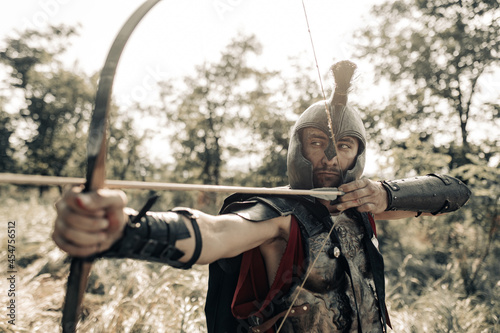 Ancient warrior archer aims and shoots from bow among meadow. photo
