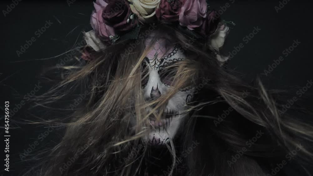 Halloween makeup, a girl in the image of a cute and good death Santa Muerte or a sugar Mexican skull calmly looks at the camera with hair in the wind in slow motion.