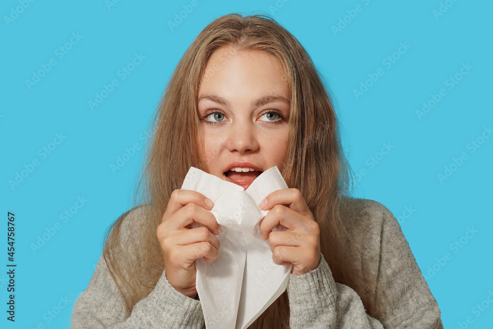Young blond woman suffering from allergy on light blue background. Symptoms of cold or allergy. Woman sneezes in white tissue. Young blond woman suffering from allergy on light blue background. Symptoms of cold or allergy. Woman sneezes in white tissue.