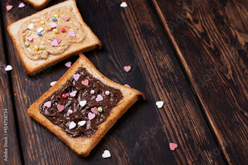 Toast with chocolate paste and peanut butter sprinkled with colorful hearts on a dark wooden background.