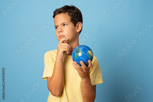 Pensive school boy holding a world globe isolated over blue background © Danko