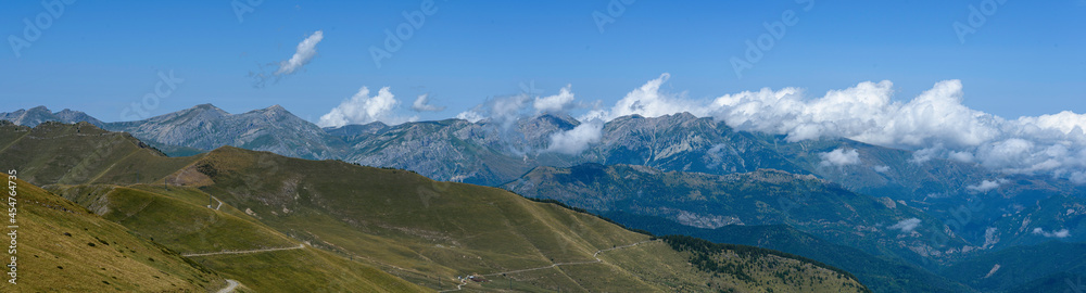 Panoramic of the peaks of the mountain range between clouds in the background, above a clear blue sky, in the foreground the hill in front without trees, with grass, grass, for cattle, and a path that