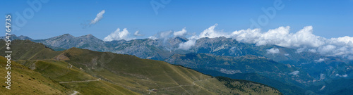 Panoramic of the peaks of the mountain range between clouds in the background  above a clear blue sky  in the foreground the hill in front without trees  with grass  grass  for cattle  and a path that