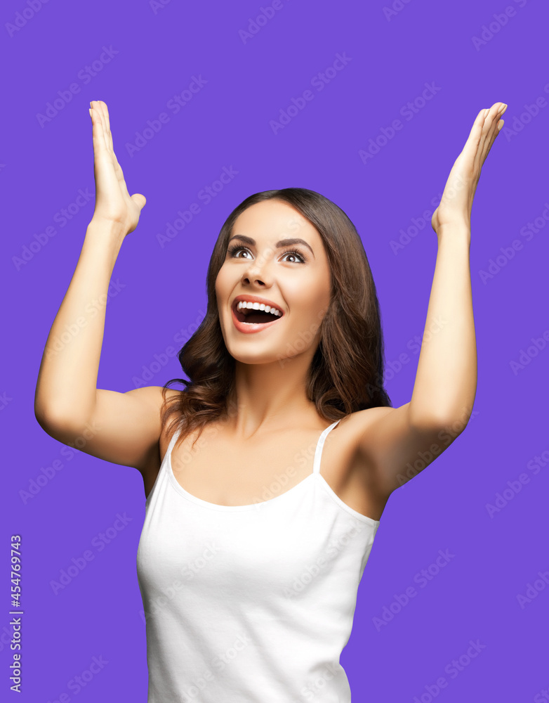 Photo of woman in white casual clothing raising hands up with flat palms, happy gesturing or praying, isolated over violet purple background. Excited brunette girl looking up with smile, at studio. Photo of woman in white casual clothing raising hands up with flat palms, happy gesturing or praying, isolated over violet purple background. Excited brunette girl looking up with smile, at studio.
