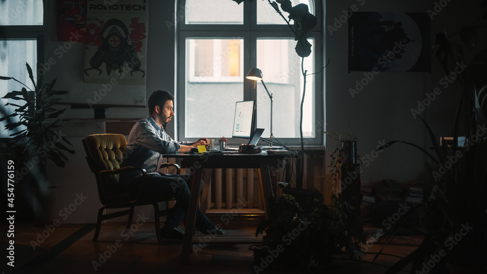 Young Handsome Man Works on a Laptop Computer in Creative Agency in Loft Office. He Writes Down a Note with Pen. Renovated Stylish Decor with House Plants, Artistic Posters and Big Rounded Windows. Young Handsome Man Works on a Laptop Computer in Creative Agency in Loft Office. He Writes Down a Note with Pen. Renovated Stylish Decor with House Plants, Artistic Posters and Big Rounded Windows.