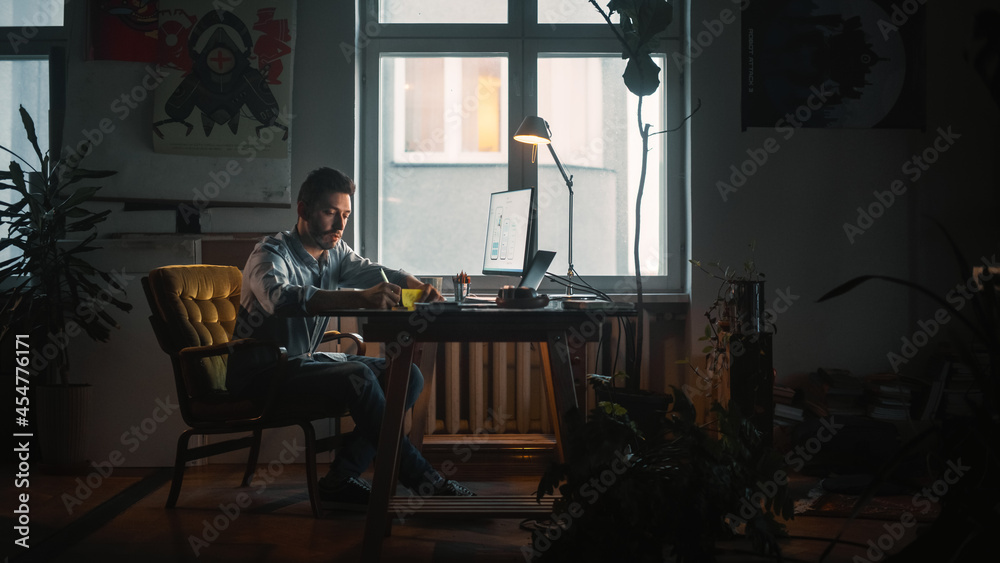 Young Handsome Man Works on a Laptop Computer in Creative Agency in Loft Office. He Writes Down a Note with Pen. Renovated Stylish Decor with House Plants, Artistic Posters and Big Rounded Windows.