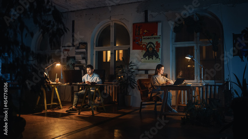 Two Stylish Employees Working on Computers in Creative Agency in Loft Office. Colleagues Answer Emails and Manage Marketing Projects. Dark Renovated Space in Evening with Plants and Artistic Posters. © Gorodenkoff
