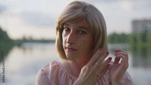 Headshot portrait of trans woman looking at camera as mirror touching hair and smiling. Close-up face of adult Caucasian transgender adjusting hairdo resting outdoors on sunny evening. Slow motion photo