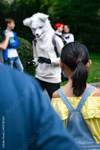 A little girl from the back in overalls looks at the furry character dressed as a white wolf or a dog with gloves. Anime parade