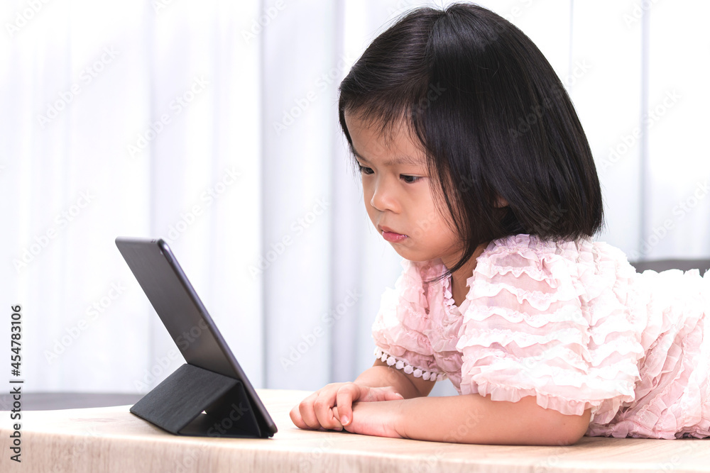 Happy child watching cartoon on tablet at living room in her house. Kid lying down on the wood table. Sweet girl wearing soft pink aged 4-5 years old. Happy child watching cartoon on tablet at living room in her house. Kid lying down on the wood table. Sweet girl wearing soft pink aged 4-5 years old.