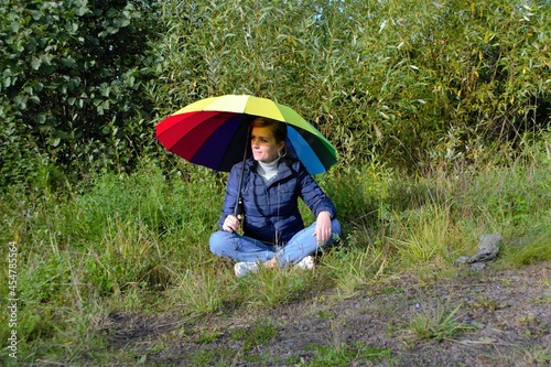 A woman in a jacket and jeans with a rainbow umbrella sits in the park on the grass. Warm autumn day © Eugenia