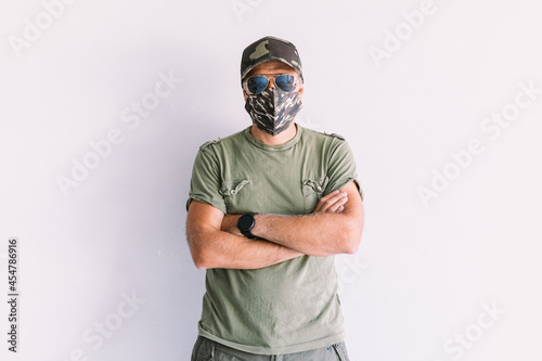 Military man with camouflage cap, sunglasses and camouflage mask, with crossed arms, on a white wall