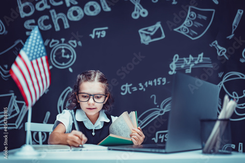 beautiful little schoolgirl sitting at desk and study online with laptop against black background with USA flag