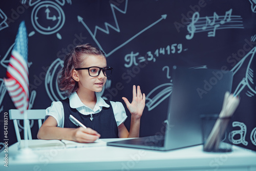 beautiful little schoolgirl sitting at desk and study online with laptop against black background with USA flag