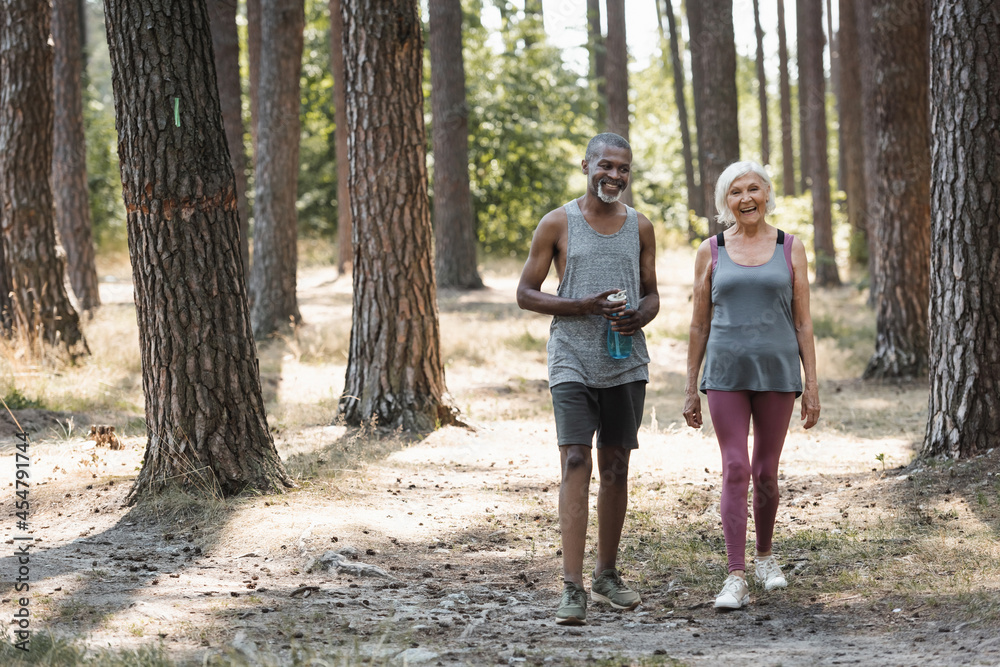 Elderly african american sportsman with sports bottle walking near smiling wife in forest. Elderly african american sportsman with sports bottle walking near smiling wife in forest.