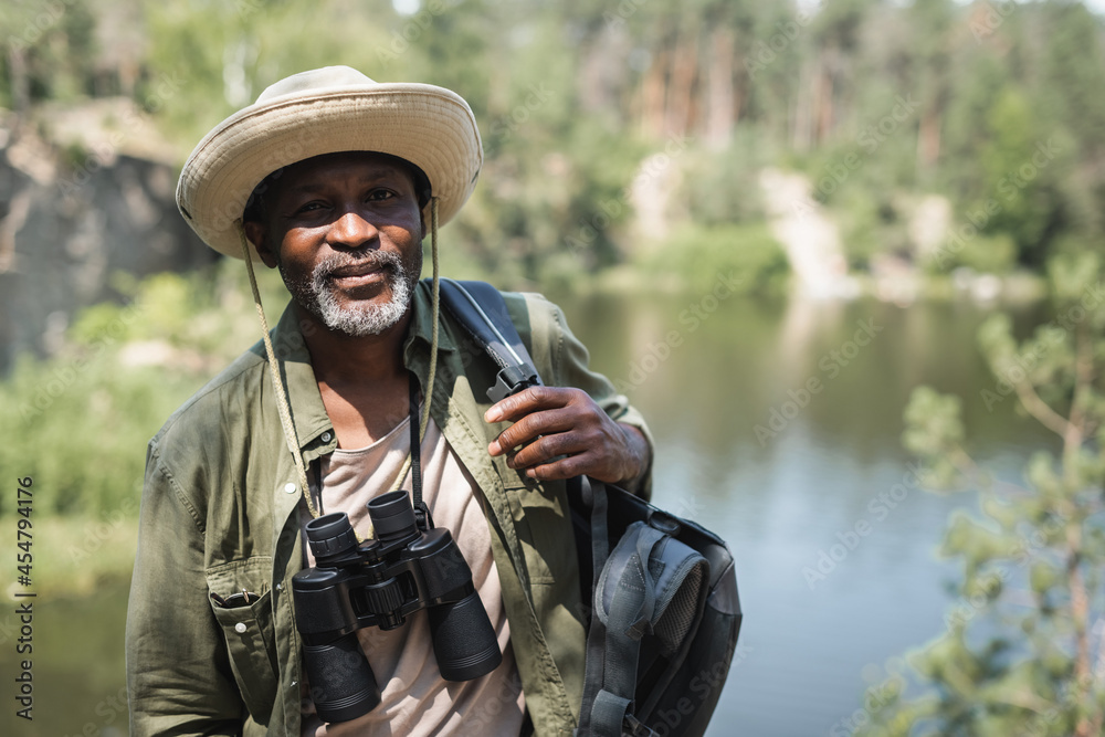 African american hiker with backpack and binoculars looking at camera outdoors.