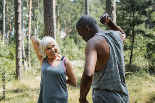 Senior woman smiling while exercising with african american husband in forest.