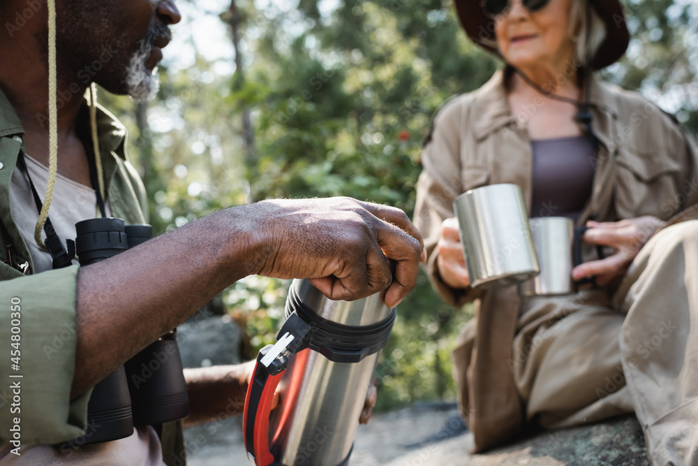 Cropped view of african american tourist holding thermos near blurred wife with cups in forest.