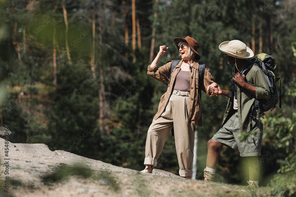 Fototapeta premium Excited elderly hiker showing yes gesture while holding hand of african american husband on rock.