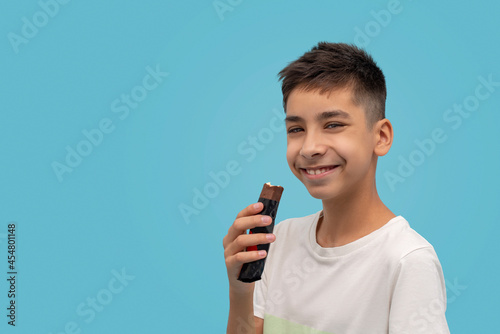 Waist up portrait of a young smiling boy teen eating chocolate against blue background in studio