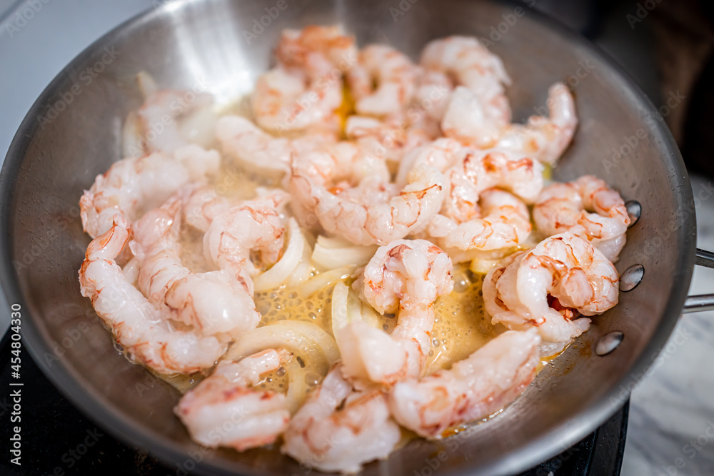 Macro closeup of stainless steel frying pan with sauteing olive oil, sweet onion slices and whole large king jumbo Argentinian shrimp seafood cooked on electric stove in home kitchen Macro closeup of stainless steel frying pan with sauteing olive oil, sweet onion slices and whole large king jumbo Argentinian shrimp seafood cooked on electric stove in home kitchen
