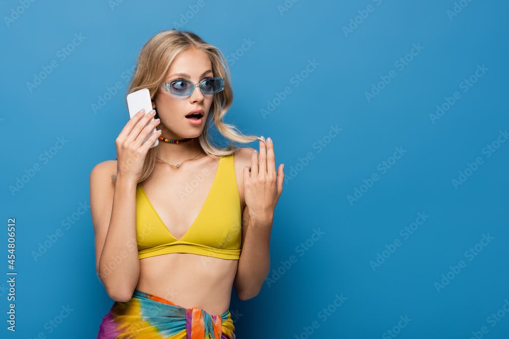 shocked young woman in yellow bikini top talking on smartphone isolated on blue.