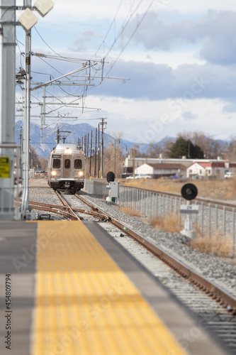 Train arriving at the station. Lights on coming down the tracks. Pedestrian platform in foreground. Rocky Mountains in background.