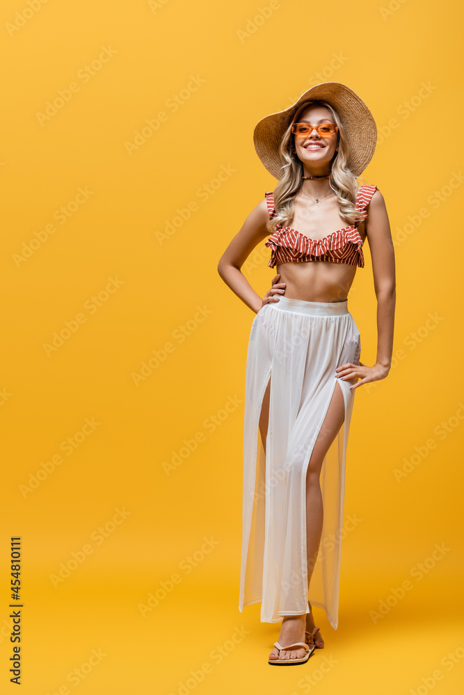 full length of smiling woman in ruffle bikini top and white skirt posing with hand on hip on yellow.